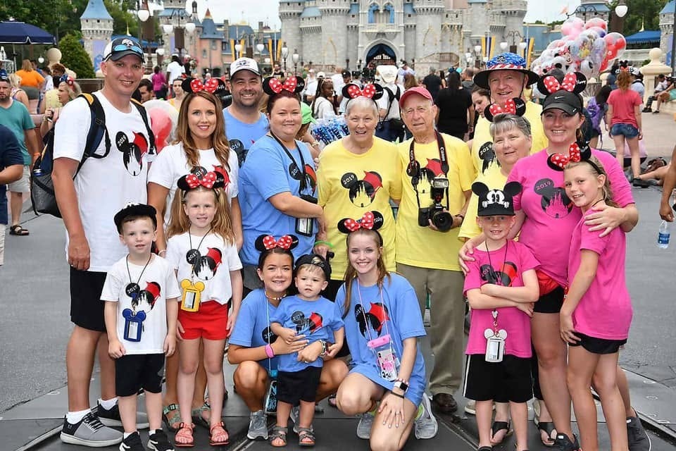 A multigenerational group in matching Disney shirts and Minnie Mouse ears poses in front of a castle.