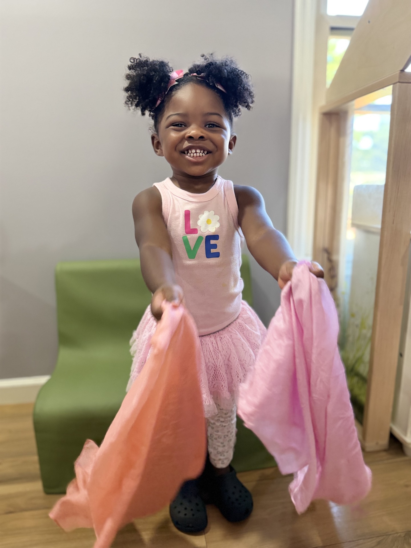 A smiling child in a pink LOVE shirt holds orange and pink play scarves beside a green chair in a classroom.