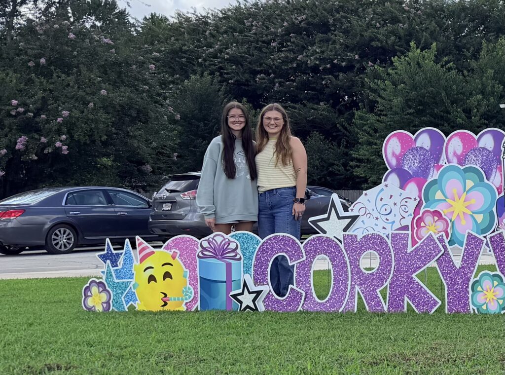 Two Corky team members standing behind a colorful Corky yard sign outside the school