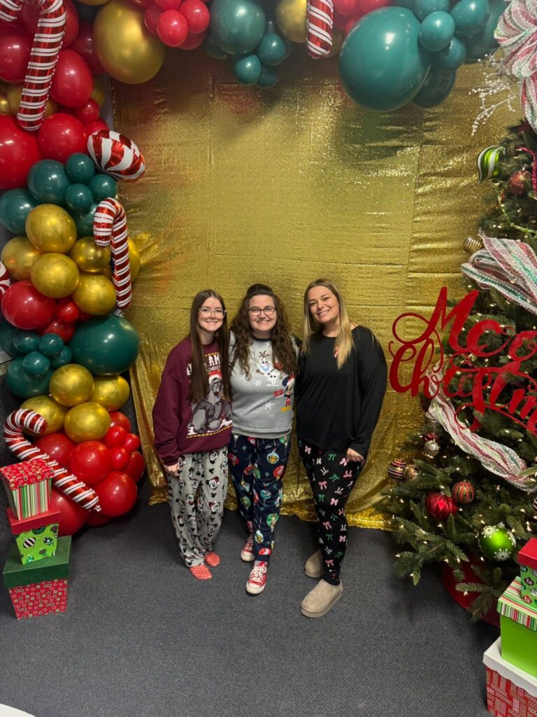 Three Corky team members in pajamas posing in front of a festive holiday balloon backdrop